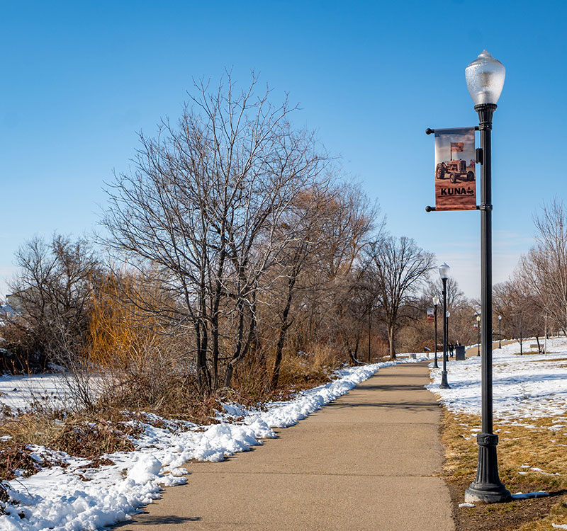 Kuna Idaho walking path along the river with a Kuna banner on a light post. The banner shows a tractor with an American flag and the words Kuna below it.