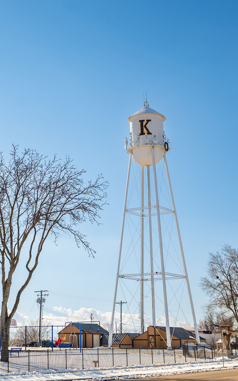 Kuna Idaho water tower. Towering high above a park with buildings in the background. The sky is bright blue. The water tower is white with the letter K on it.
