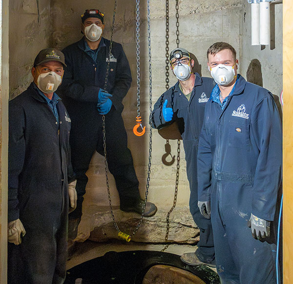 Group of 4 plumbers in a sewage well with overalls and masks on.